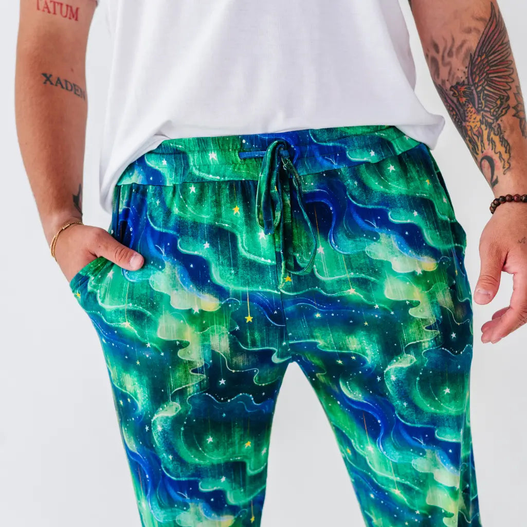 Close-up of pajama pants with a swirling green and blue northern lights pattern, showing the drawstring waist and a hand in the pocket.